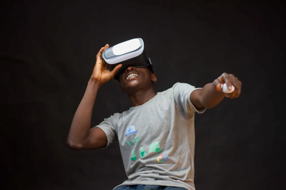 person wearing grey shirt and VR glasses, against black background. The person is leaning back, smiling, and gesturing forward with their hand, which is holding a remote control type of device