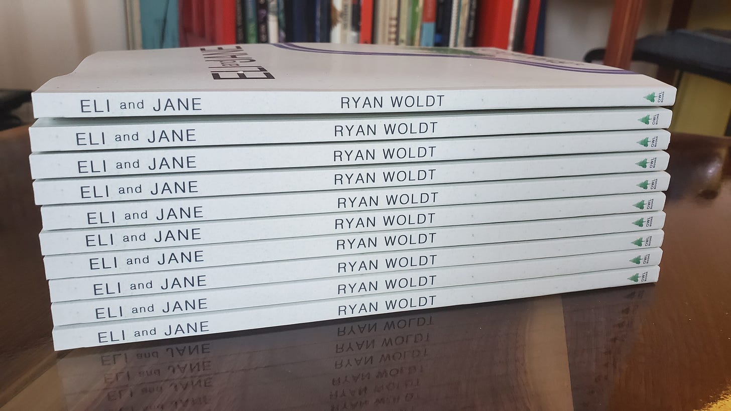 10 copies of the book "Eli and Jane" by author Ryan Woldt sitting on a wood coffee table. Book is white, and font is sans serif. Close up view of the spine.