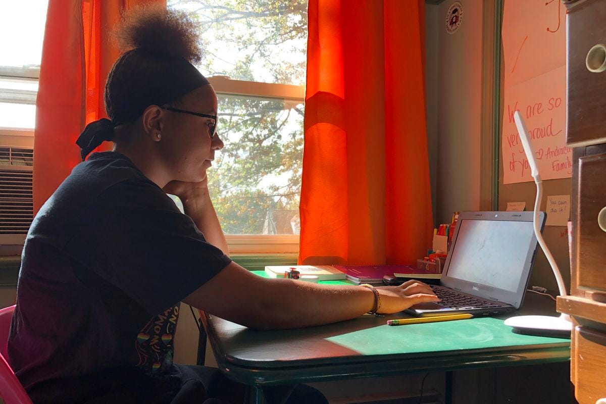 A teenage girl participates in a remote class on her laptop, sitting at a desk in her room.
