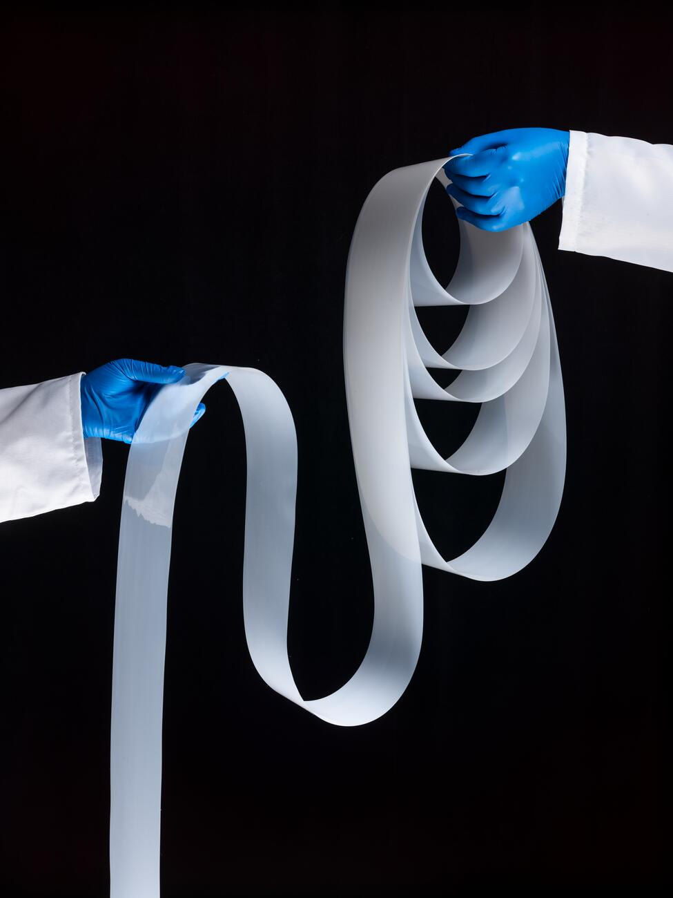 Picture of white ribbon held by two hands in blue gloves on black background