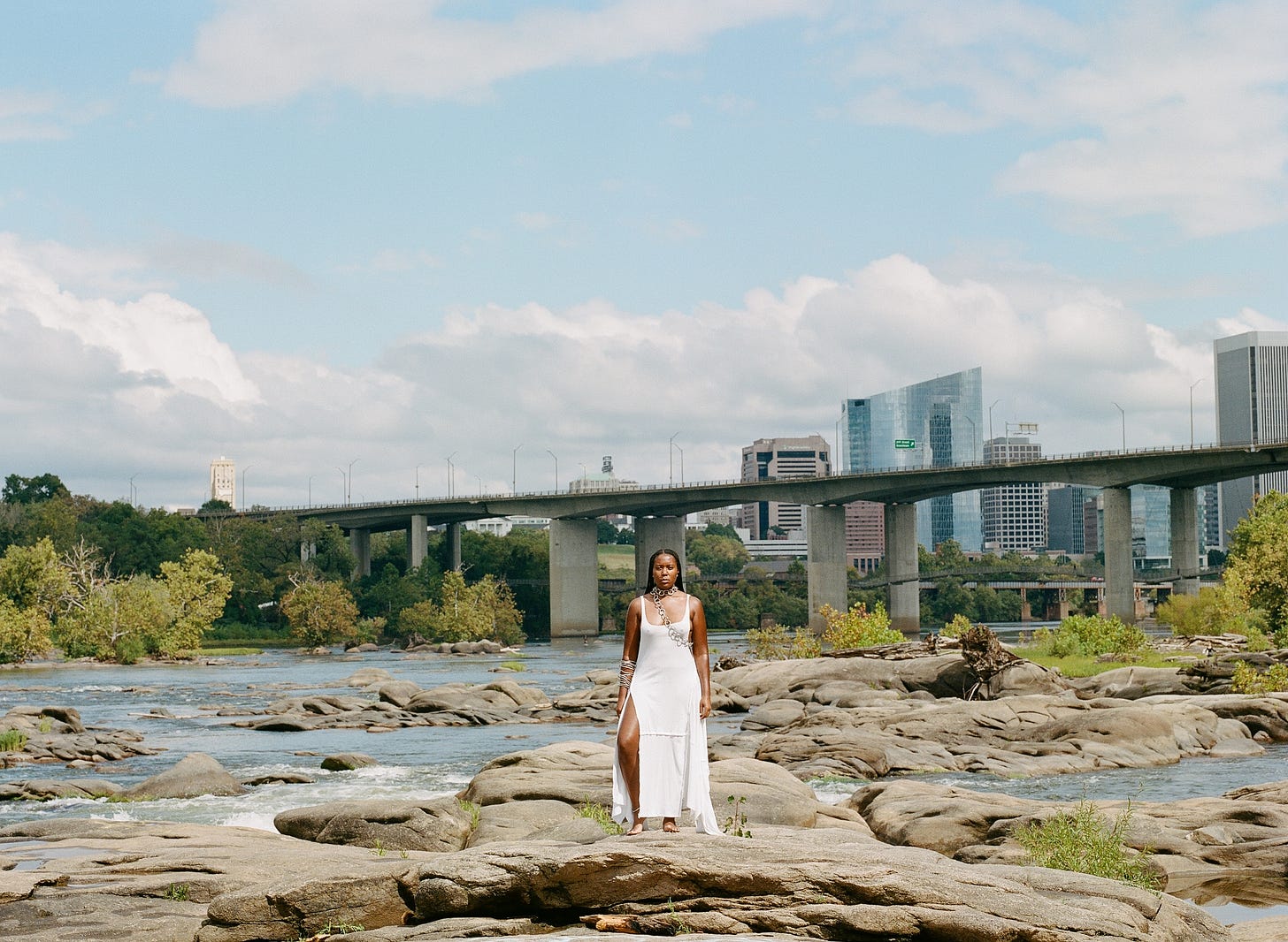 A black woman (me) with braids wearing a white dress looking into the camera while standing on top of river rocks with the city line in the background.