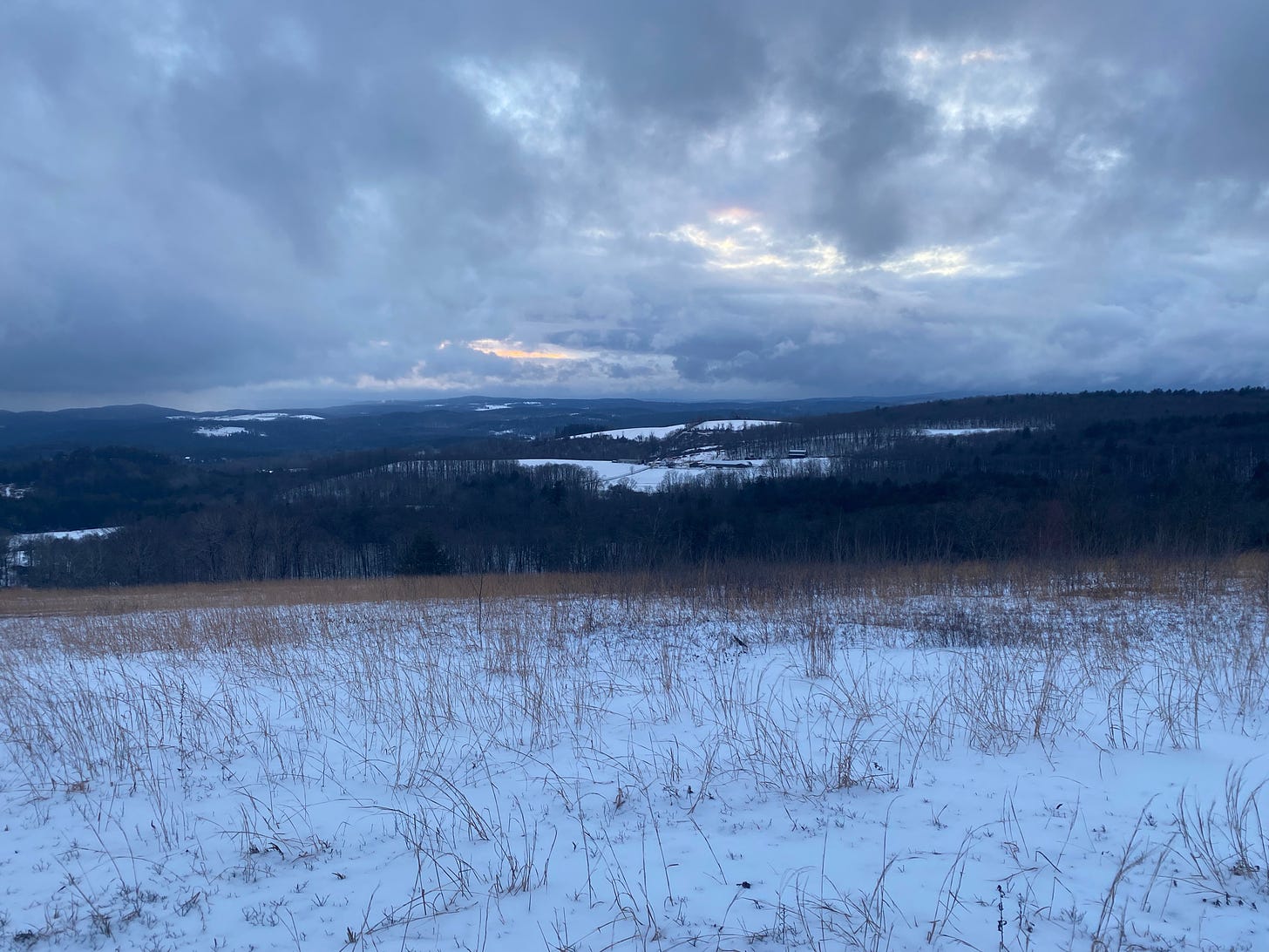 A snowy ridgetop looking out over a view of dark forested hills and snow-covered fields. The sky is dark, full of big blue and purple clouds, and a small slice of orange light where the sun is peeking out.