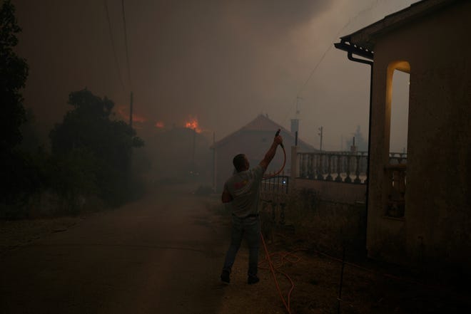 An man uses a garden hose to water the roof of a house as a forest fire smoke darkens the sky in the village of Bemposta, near Ansiao, central Portugal, Wednesday, July 13, 2022. Thousands of firefighters in Portugal continue to battle fires all over the country that forced the evacuation of dozens of people from their homes.