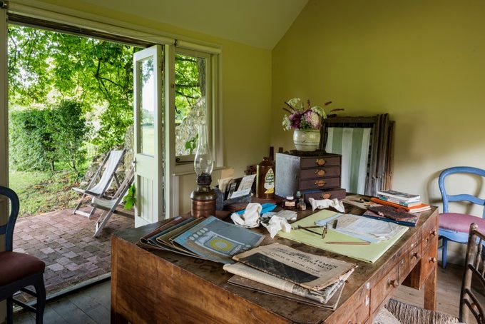 A view from behind the desk in the writing lodge at Monk's House. Papers, pencils and a discarded pair of glasses lie strewn across the desk. Beyond the open door of the lodge, two deckchairs can be seen on a patio shaded by trees.