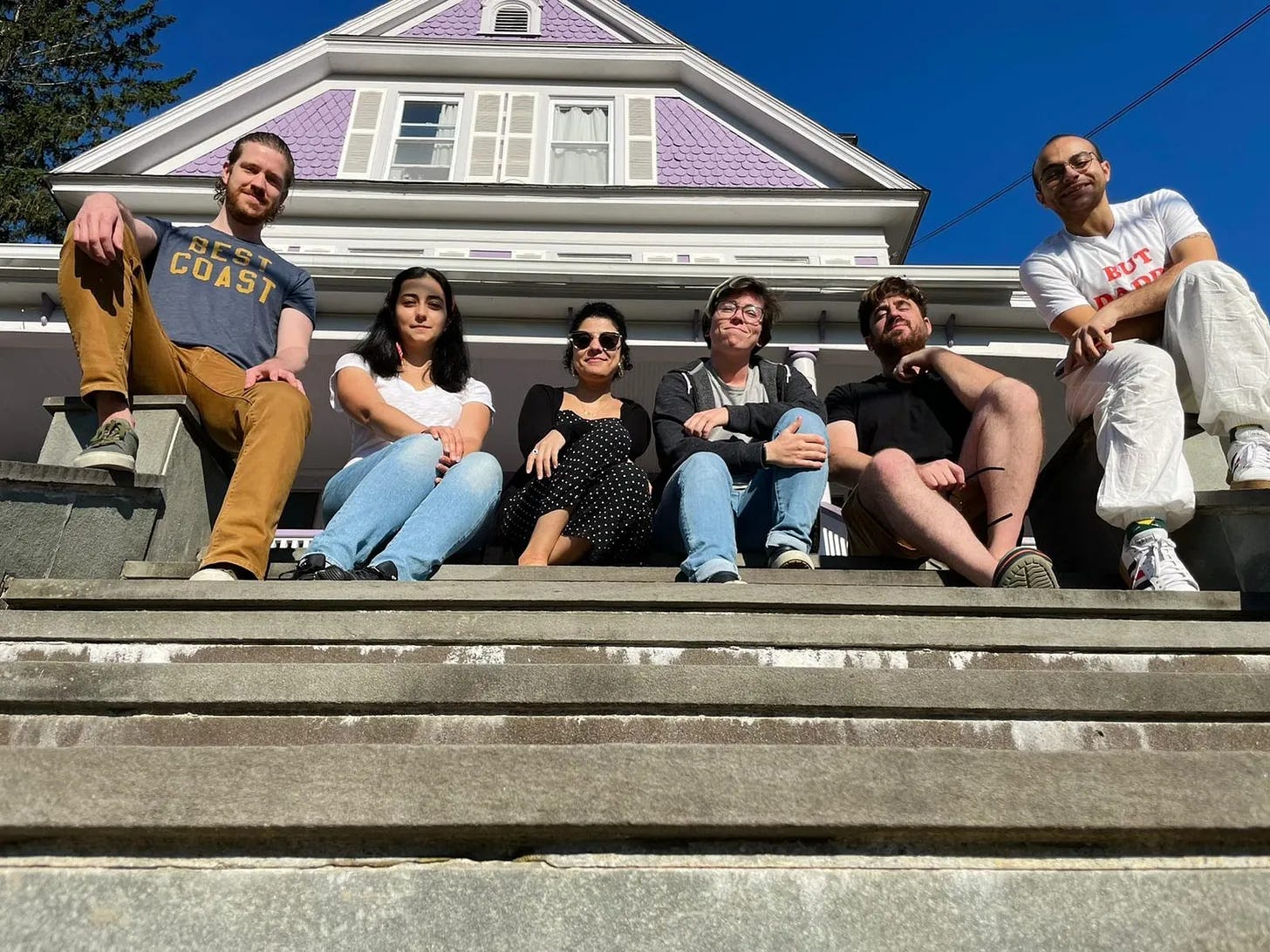 Six young people sit on the stairs of a three story purple house, looking down at the camera