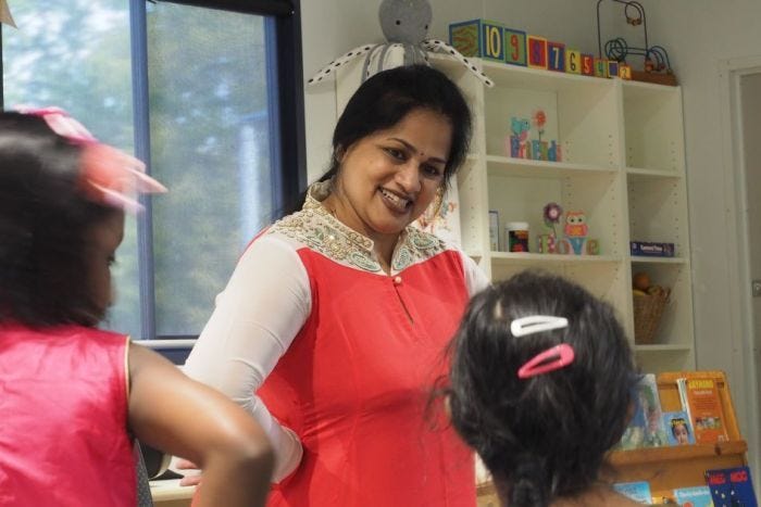 An Indian dance teacher stands with hands on hips in a children's classroom and smiles at two young female students.
