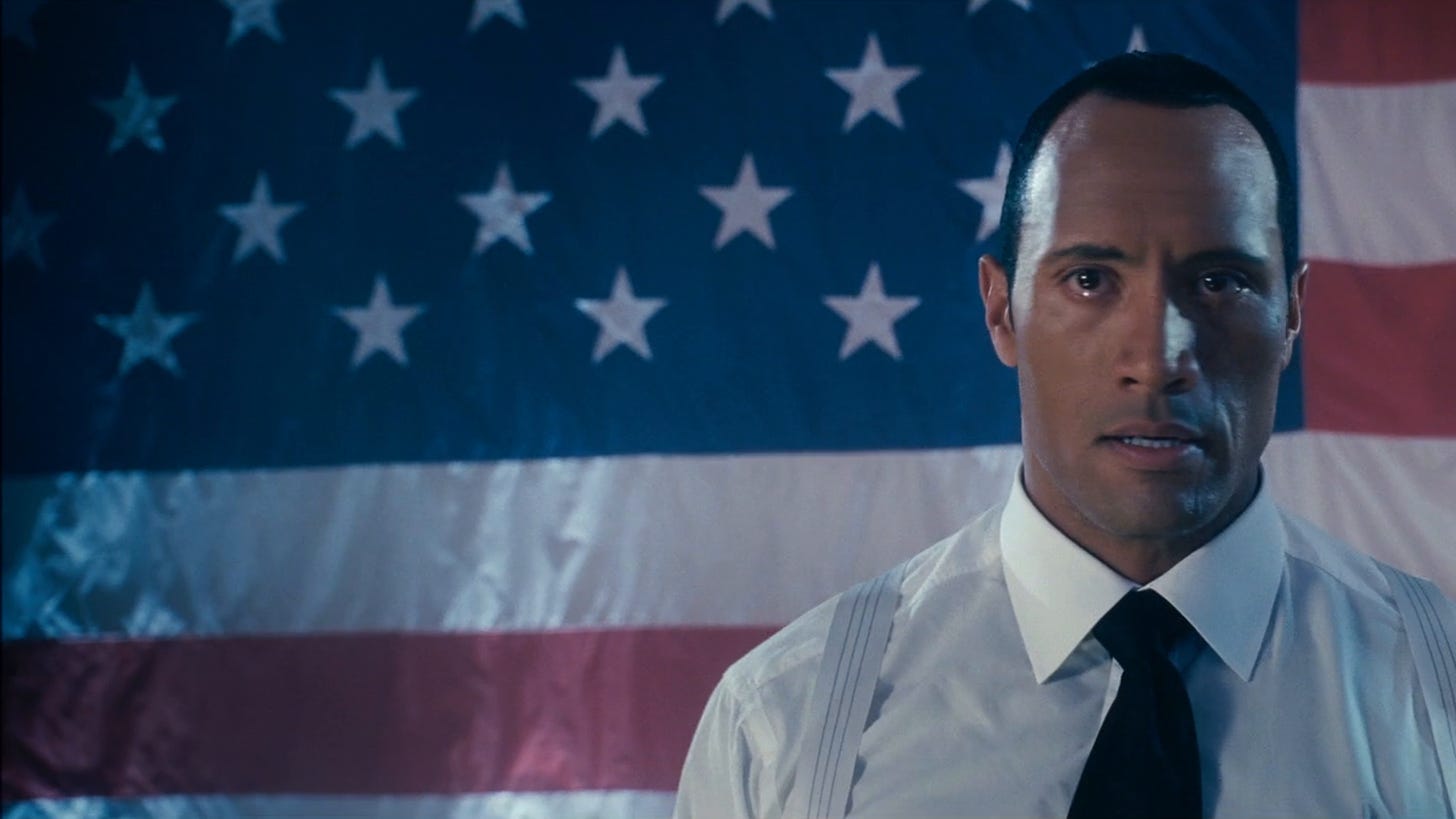 Movie still from Southland Tales. The Rock is in a white dress shirt and tie, standing in front of the American flag with tears in his eyes.