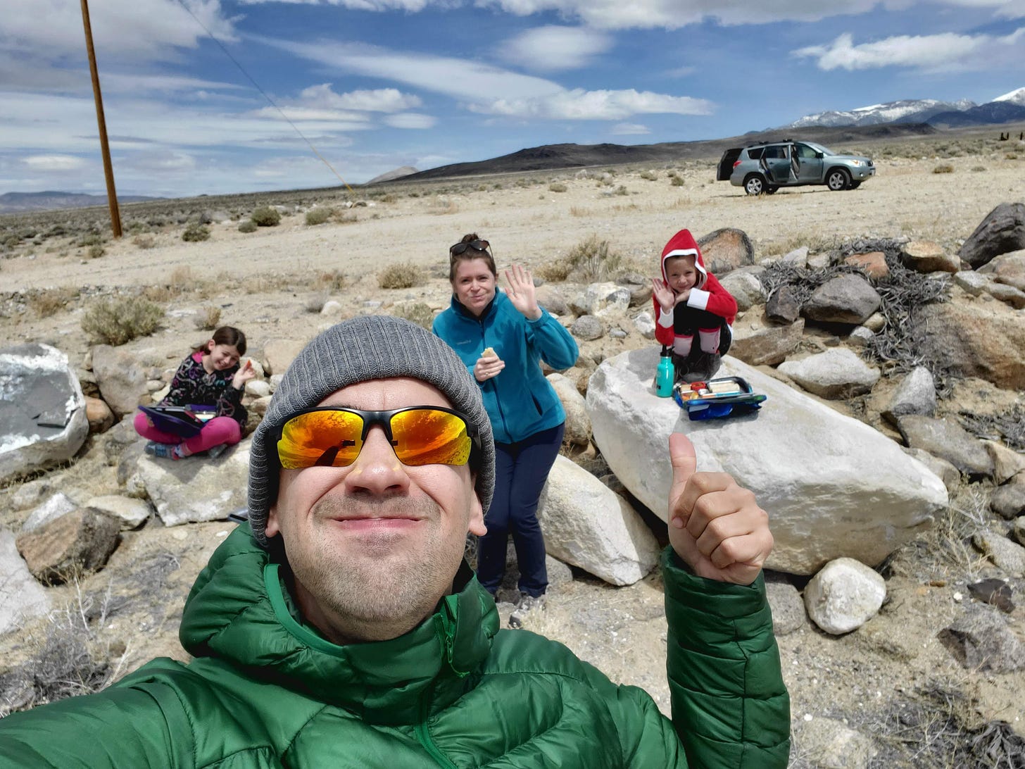 Eric taking a selfie with C, Charlotte, and L behind him sitting on rocks with their lunches in front of them.