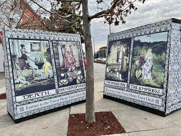A photo of two large freestanding walls on a street corner that have giant tarot cards wheat-pasted onto them as an installation for something called Journey to the Tower Photo Tarot Deck, by artists Two by Sea. These are a photographic interpretation of the classic Rider Smith Waite deck. In this photo, you can see Death and The Hierophant on one wall, and The Magician and The Empress on the other.
