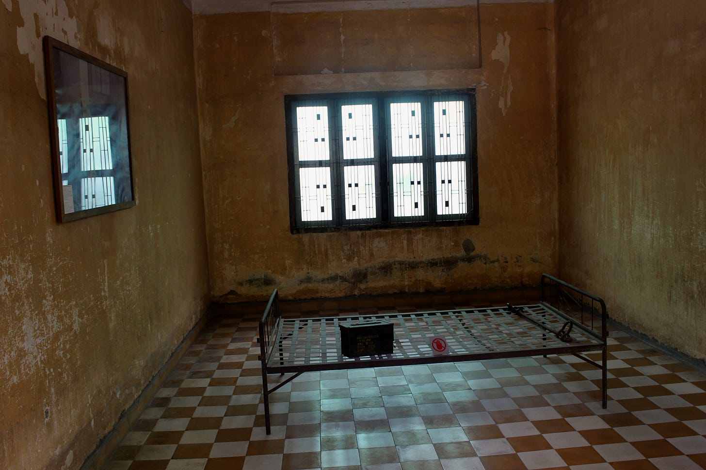 A room where the walls and floor are faded and worn. There is a single bedframe in the center of the room, as well as a single mirror and some windows.