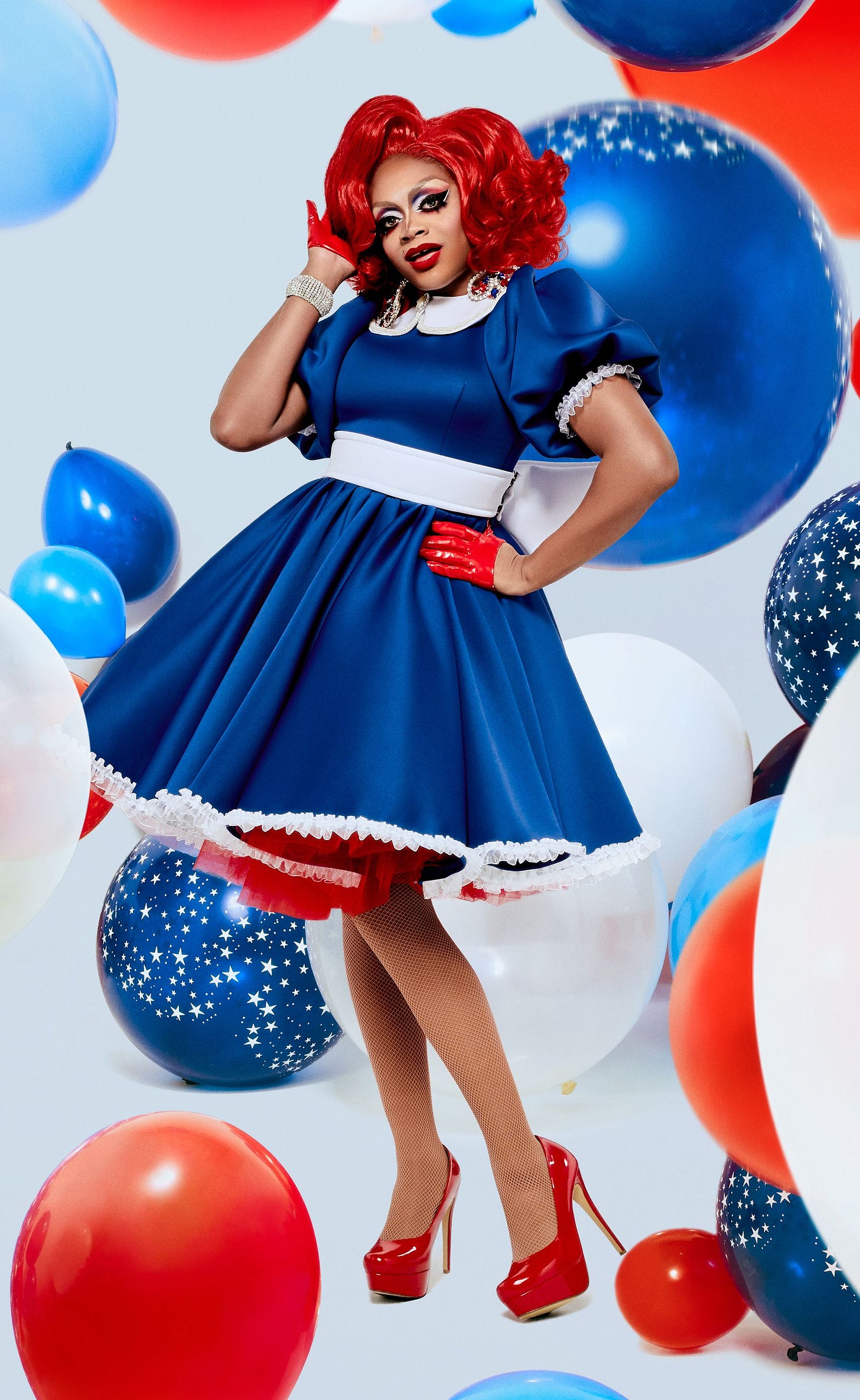 A staged promotional photo of Heidi N Closet. She wears a red wig, a blue dress with puffy sleeves and a white sash with red high heels. The effect is very reminiscent of the American flag. She poses with one hand on her hip and the other in her hair, with a confident expression on her face.