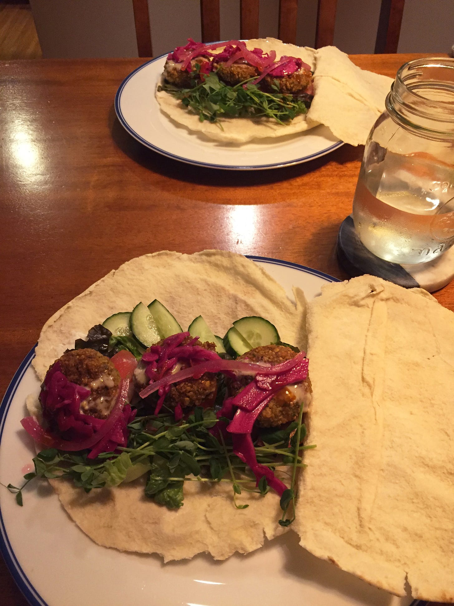 Two plates across from each other, each with an open pita full of vegetables, pink pickled onions & cabbage, three falafel patties, and a drizzle of tahini sauce.