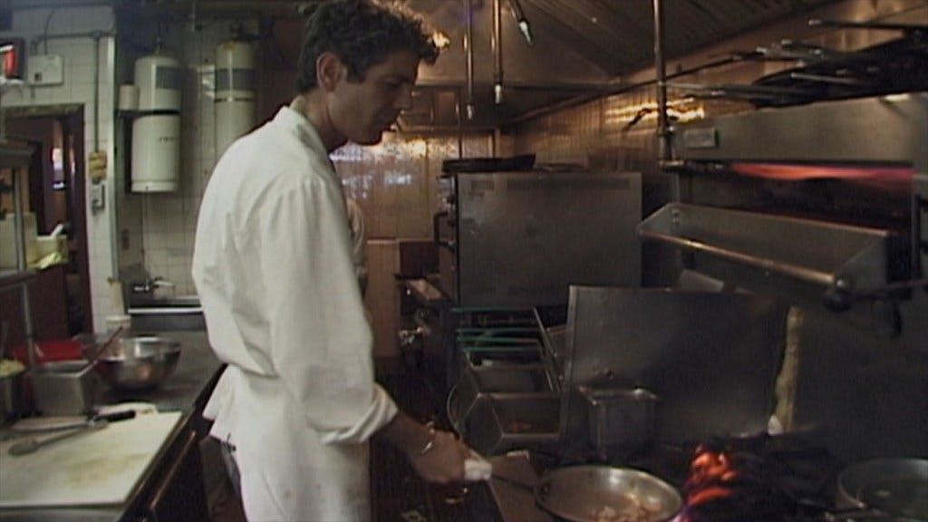 A still from Roadrunner. A young Anthony Bourdain stands in a kitchen, cooking something in a pan.
