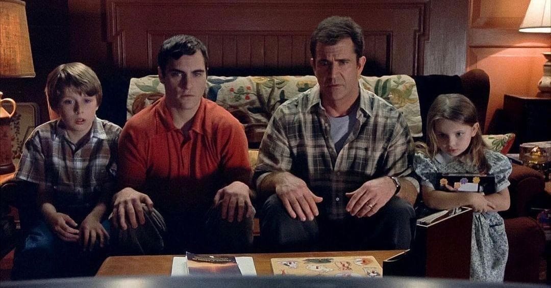The four members of the Hess family sit on the living room couch looking at the TV, which is just below the camera angle. They each look horrified and transfixed, and have their hands placed on their legs.