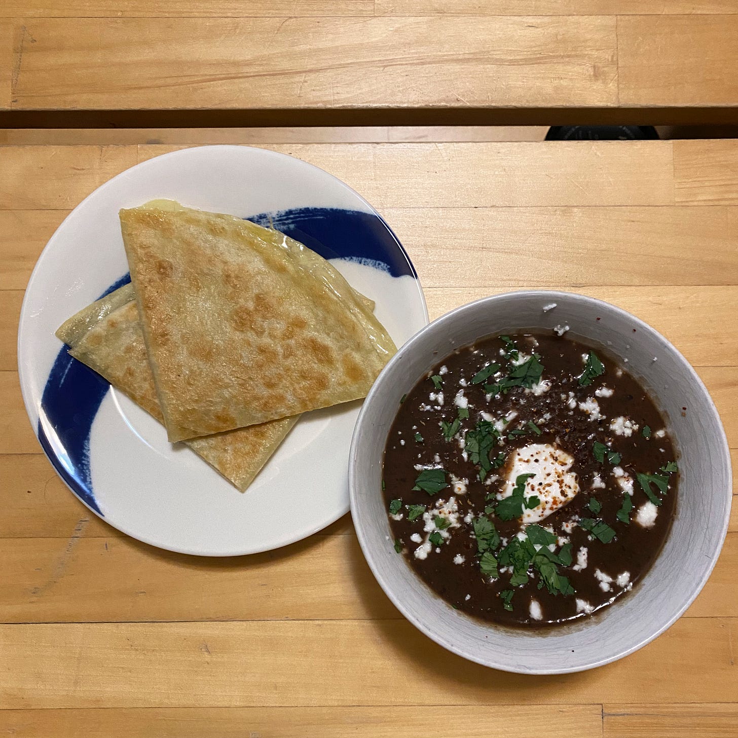 A small white bowl of black bean soup with sour cream, cilantro, feta, and tajin on top. Next to it is a white and blue plate with two quarters of a quesadilla stacked on top of each other.