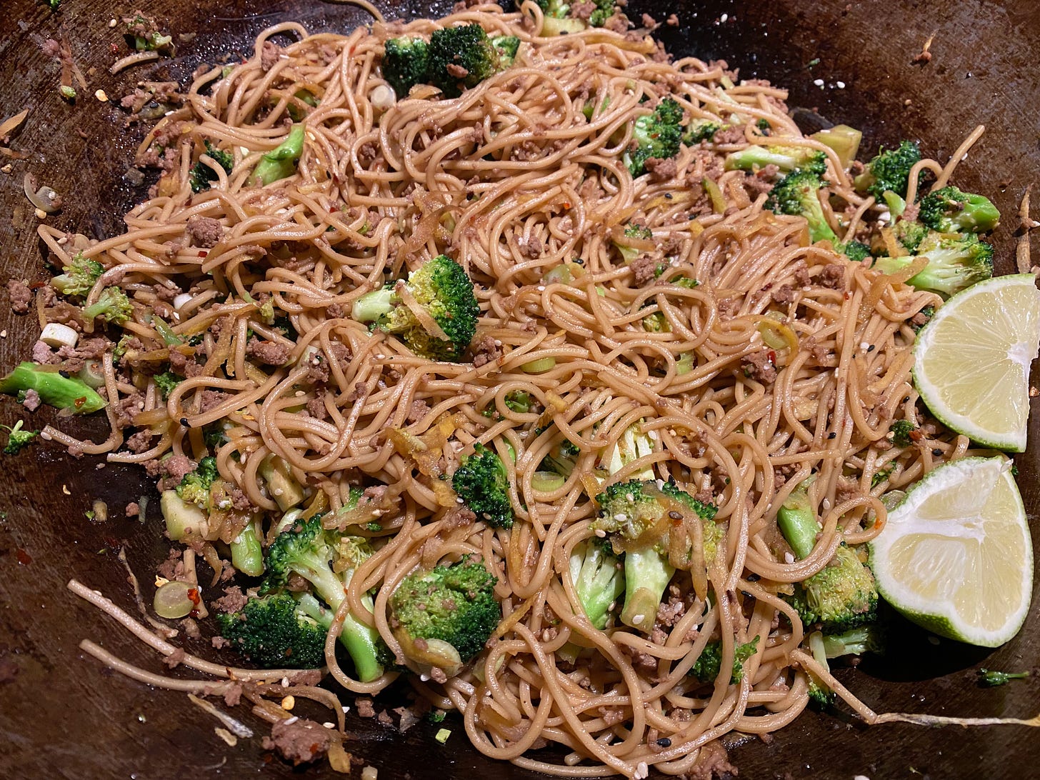 Close up of a pile of Lo mein noodles, ground pork, and broccoli florets in a wok. Two slices of lime are visible on the edge of the photo.