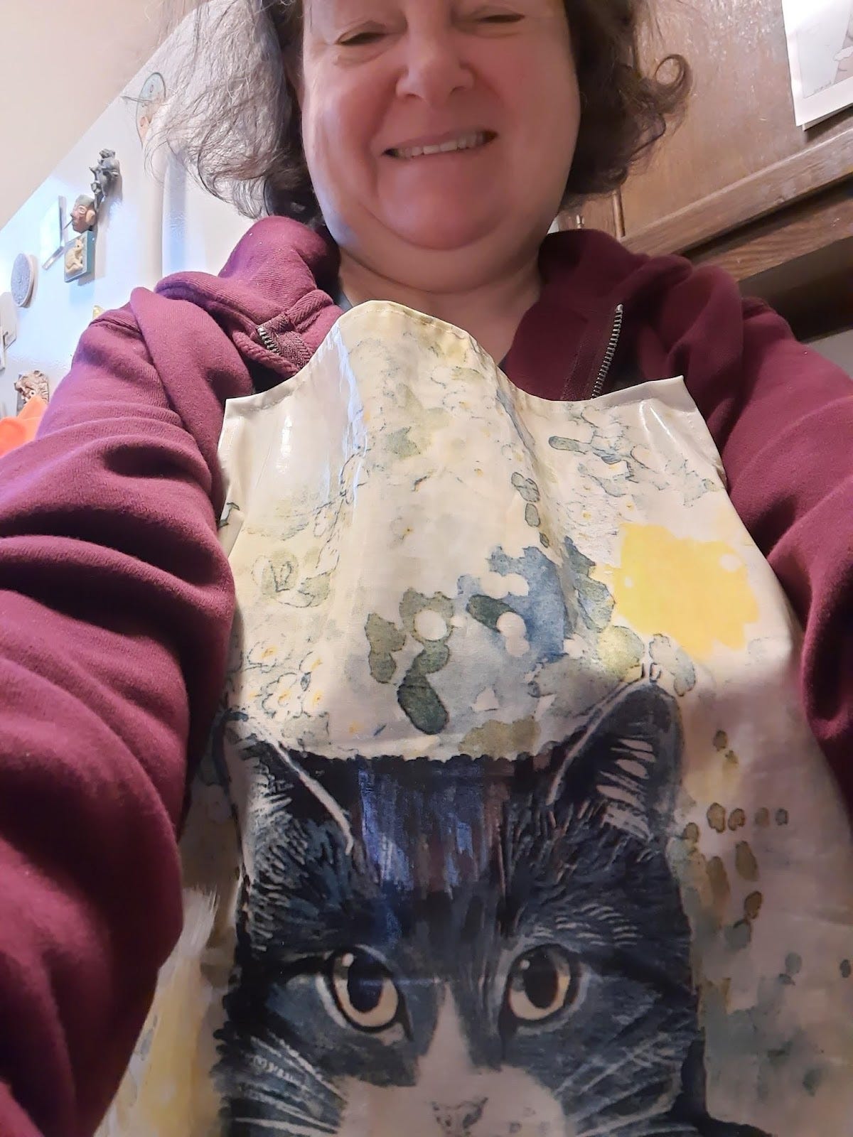 The author in her apron with a cat on it. The cat is black and white. The author is white with brown messy hair and is giggling because her arms were too short to take the photo she wanted to.