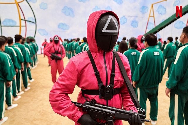 Guard in pink jumpsuit holding gun patrols single file lines of players in green jumpsuits in a still from Squid Game