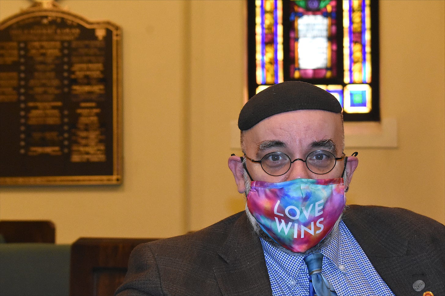 A white man in a kippeh and tye-dyed facemask that reads "Love Wins" sits in the chapel of a synagogue.) 