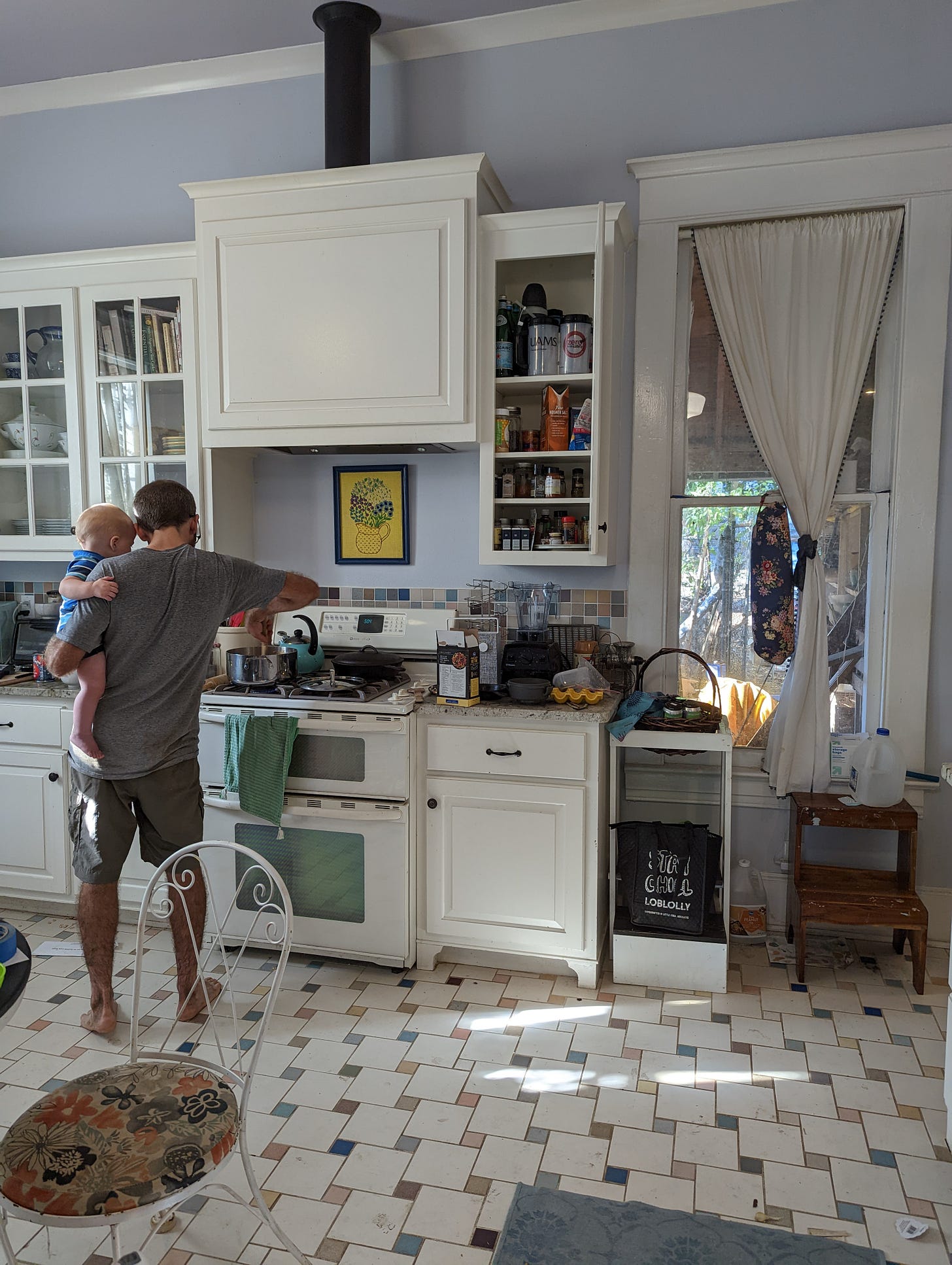 A man holds a baby while cooking with his back to the camera in a kitchen with white glass-front cabinets, lots of natural light, and a colorful checked floor tile