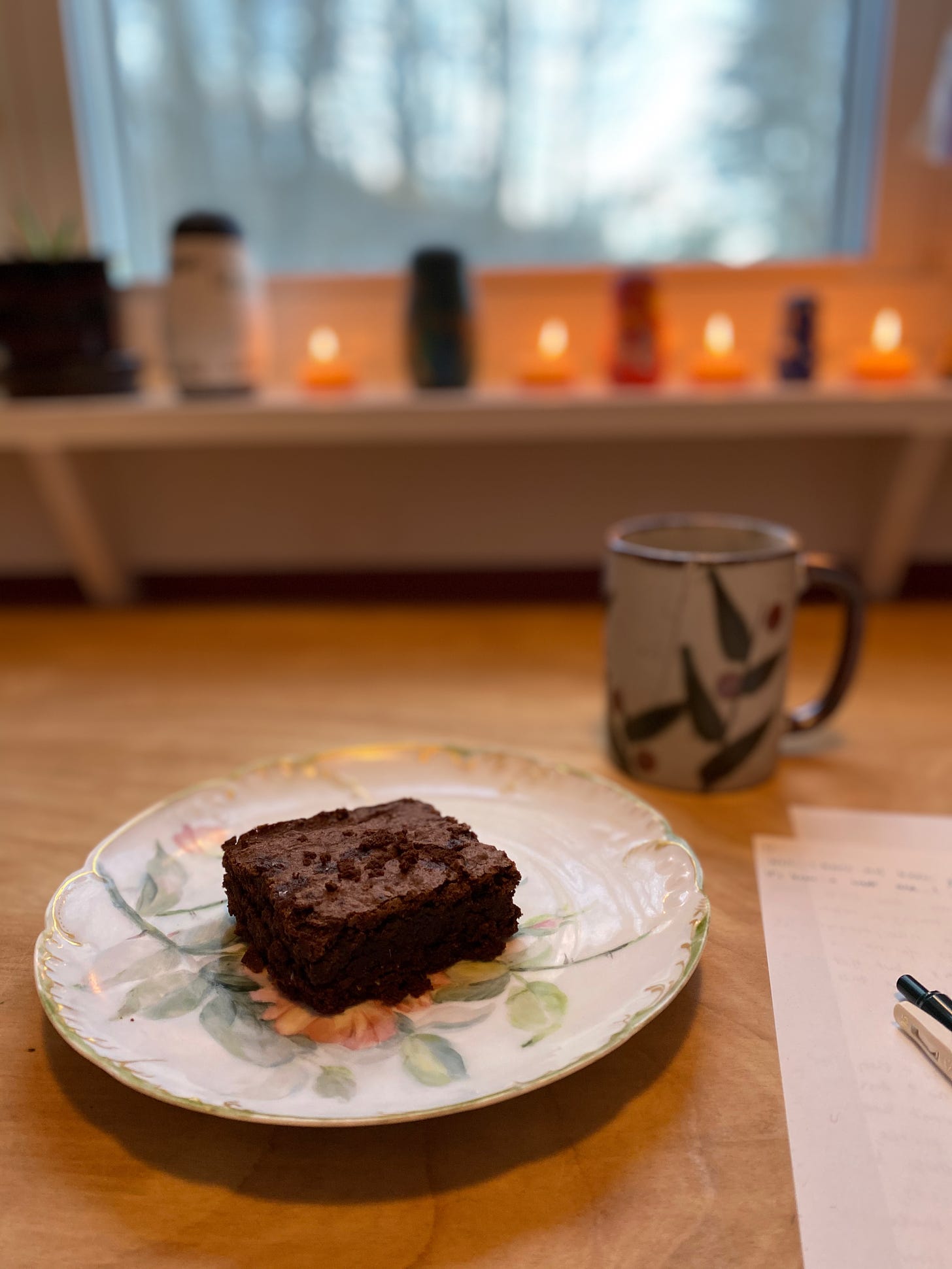 A brownie sits on a painted china plate on a wooden desk. There is a mug of tea behind the plate, and a few papers and a pen rest on the desk. In the background, there is a widow with a view of blurry trees; small lit candles and a series of painted wooden nesting dolls sit on the windowsill.