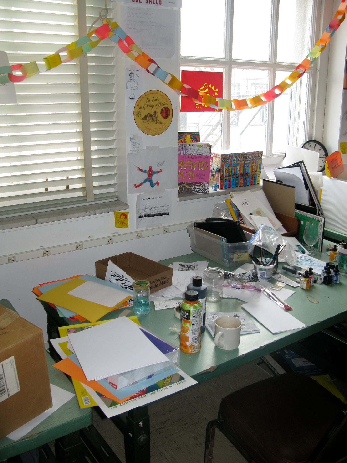 A messy desk, covered with bottles of ink, books, and other art supplies. Lots of colorful decorations are throughout.