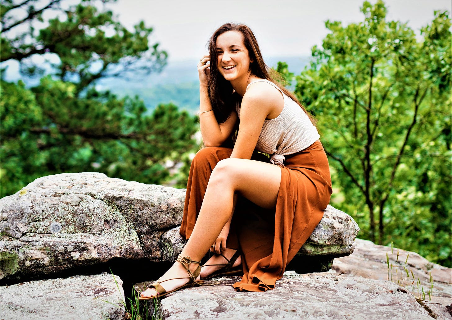 smiling girl with long brown hair sitting on boulder with trees and valley behind her