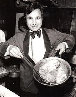 A black-and-white photo of Mr. Aslam in a tuxedo, smiling and holding up a frying pan with chicken cutlets in it.