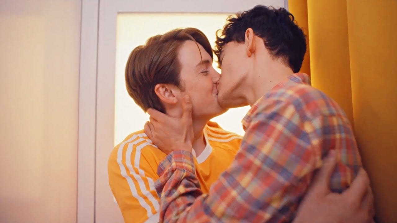 Nick, a blonde cis teen male, and Charlie, a brunette curly haired teen male, hold each other and kiss inside of a photo booth