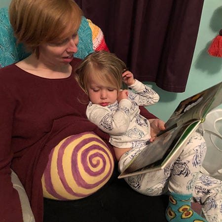 Me sitting in a chair reading a book to my kid who is in my lap staring at my pregnant belly painted in a yellow purple swirl instead of the book.