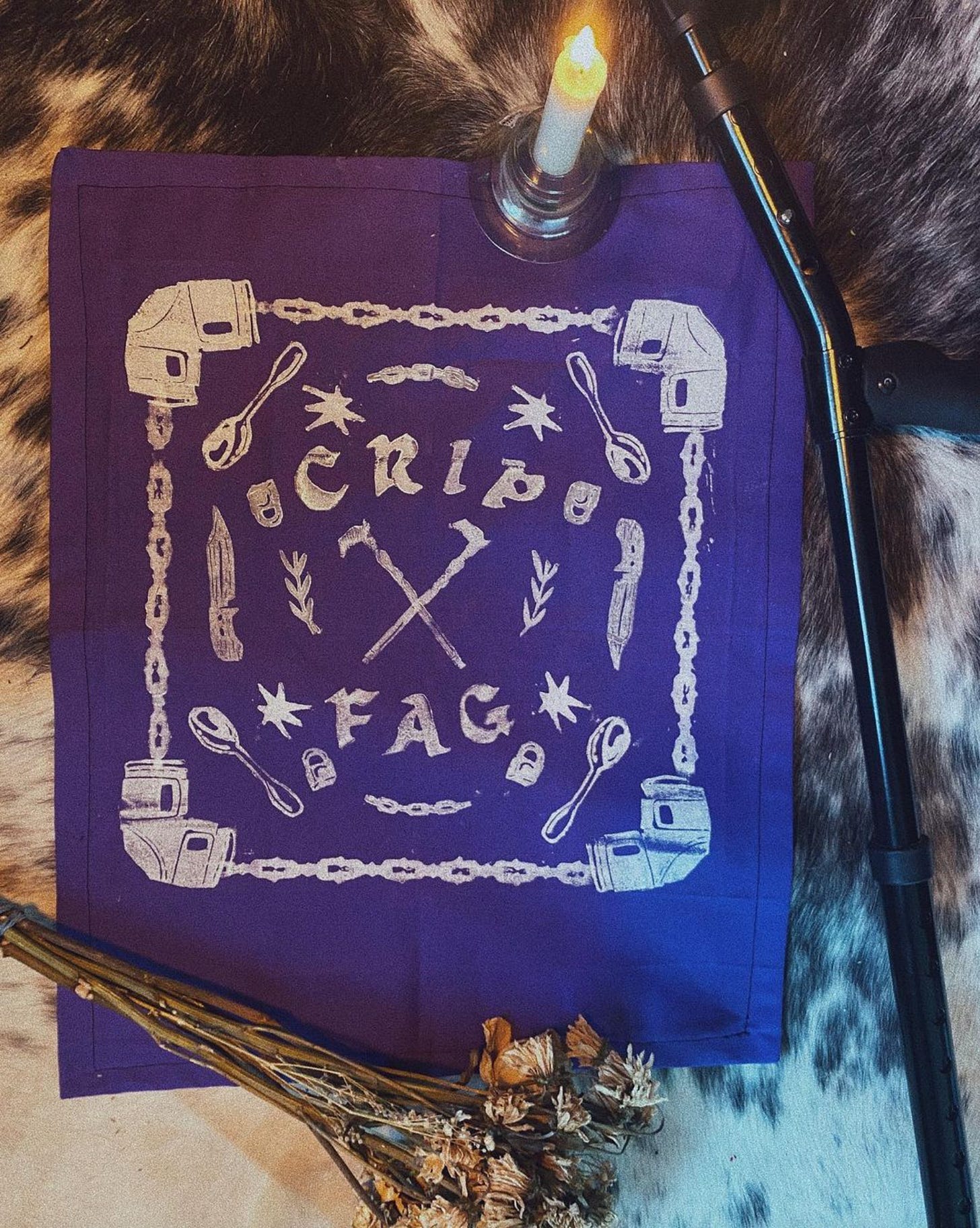 purple “crip fag” hanky or fabric hanging, laying flat on a cowhide surrounded by a taper candle, dried flowers, and a forearm crutch. the design on the hanky has a centre piece of two crossed derby-handle canes, framed by two sprigs of leaves, and the words “crip fag” above and below. Around the outside of the centre are stamps of pocket knives, padlocks, spoons, and chains. The border of the design is made of a chain pattern, with a knee/elbow brace in each corner.