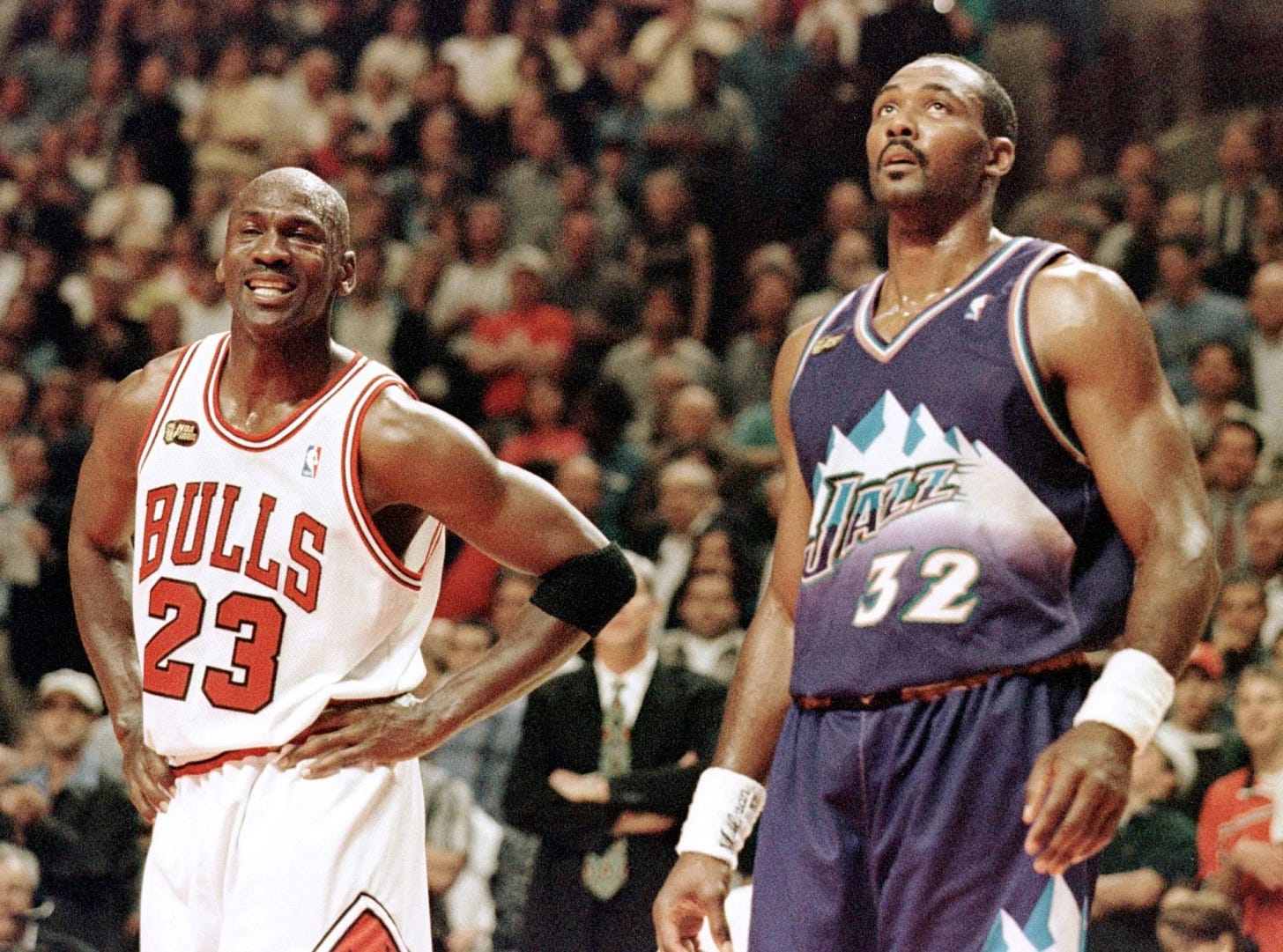 In 1998, a smiling Michael Jordan in a white Bulls uniform, hands on hips, stands beside a concerned-looking Karl Malone in a purple Jazz uniform, against the backdrop of blurry fans in arena seating.