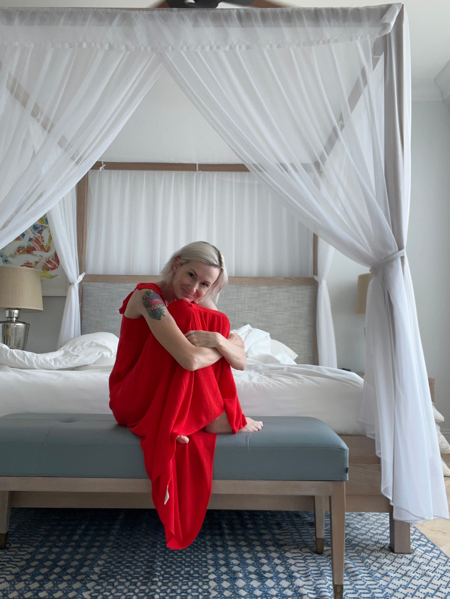 A white woman with blonde, chin-length hair, dark eyes, and a colorful tattoo on her left arm, sits in a bright red dress, knees pulled up to her chin and feet bare, on a bench in front of a white, four poster unmade bed, drapped with sheer white fabrics. 