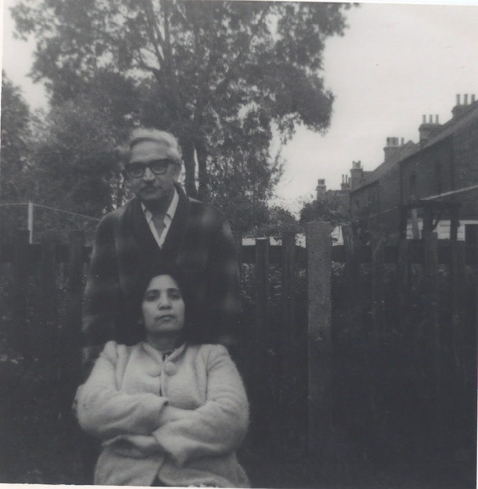 A medium sized black and white photo of two people. The man is wearing glasses and standing. The woman has her arms crossed and is sitting down