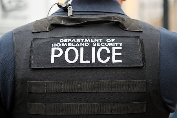 The view of the back of a police officer who is wearing a bullet proof vest that says "Department of Homeland Security: Police. 