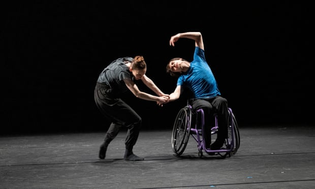 Kristen McNally and Joe Powell. Two white dancers on a black stage, one using a manual wheelchair, arching like the letter C in the YMCA dance, the other grabbing his arm, hunched over.