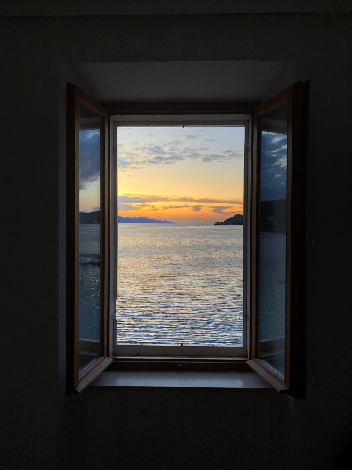 An open window that overlooks a calm sea with the sun setting across the horizon