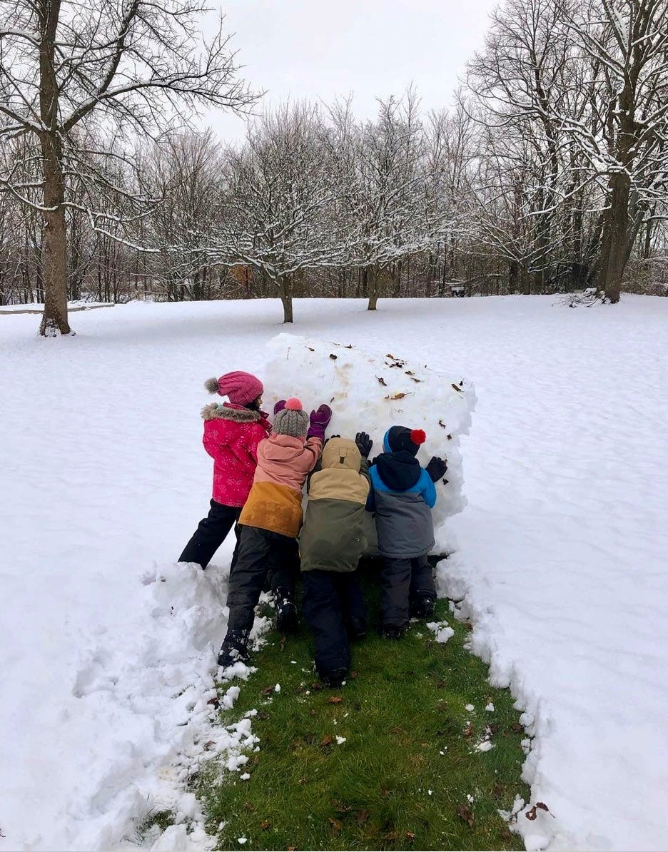 children rolling up a big roll of snow, exposing grass on the ground