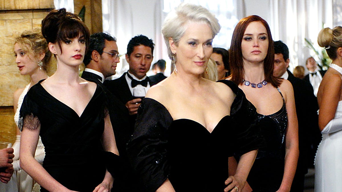 Three white women wearing gowns, portrayed by Anne Hathaway, Meryl Streep, and Emily Blunt, stand at a gala