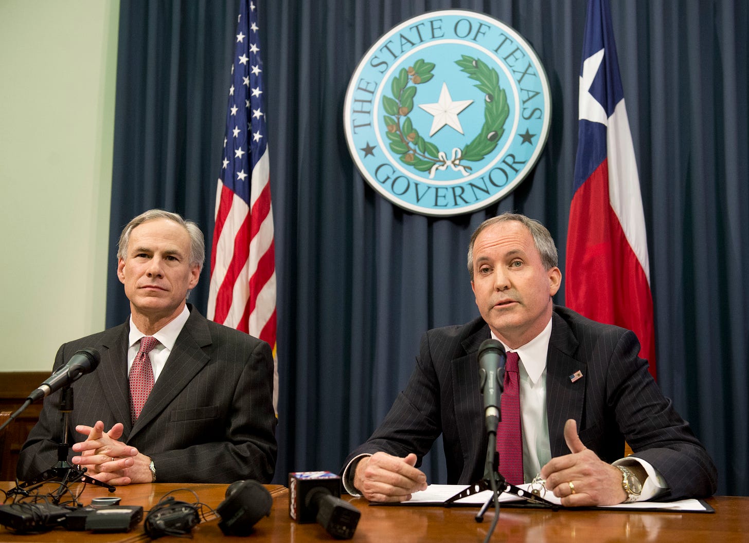 Texas Gov. Greg Abbott, l, and Attorney General Ken Paxton hold a press conference, seated at a brown desk in front of microphones, with the Texas and U.S. flag behind them, as well as the governor's seal.