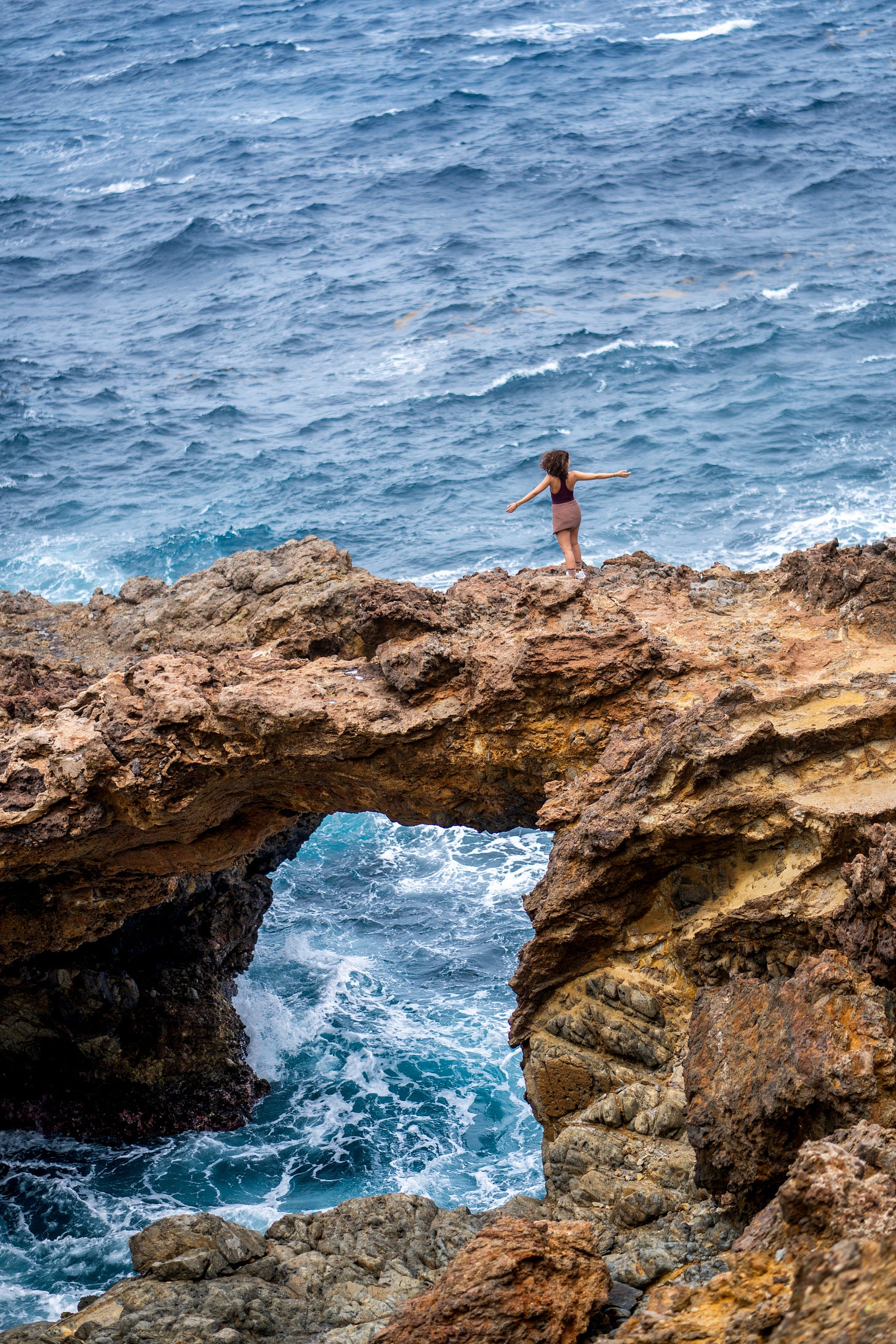 Jennifer Elrod stands on a natural cliff called the Seroe Colorado Cliffs in Aruba. Her arms are spread wide as the ocean churns beneath her.