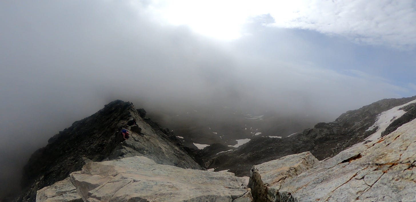 Clouds roll in over the narrow rock ledge. Two climbers straddle the rocks and scoot across toward the summit.