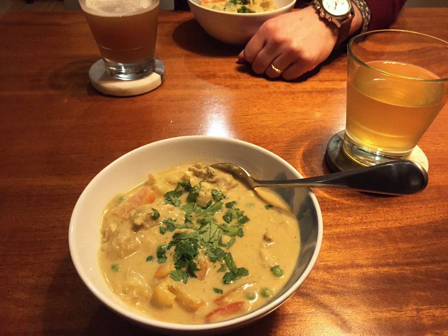 A white bowl full of curry, topped with chopped cilantro. A glass of beer sits on a coaster next to the bowl, and Jeff's hand is visible next to his bowl on the other side of the table.