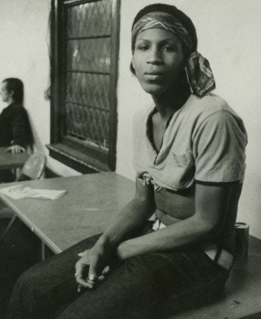 Zazu Nova, a Black trans woman, sits on a table beside a window at a meeting for the Gay Liberation front. She wears a crop top, headband, blue jeans, and a white belt.