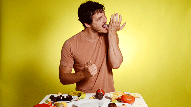 gif animado do ator Pedro Pascal lambendo a mão lambuzada de frutas — há várias diante da mesa diante dele