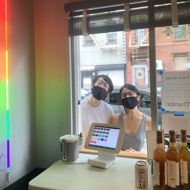 A man and woman wearing black masks in a shop window, with a rainbow-colored light along the side.