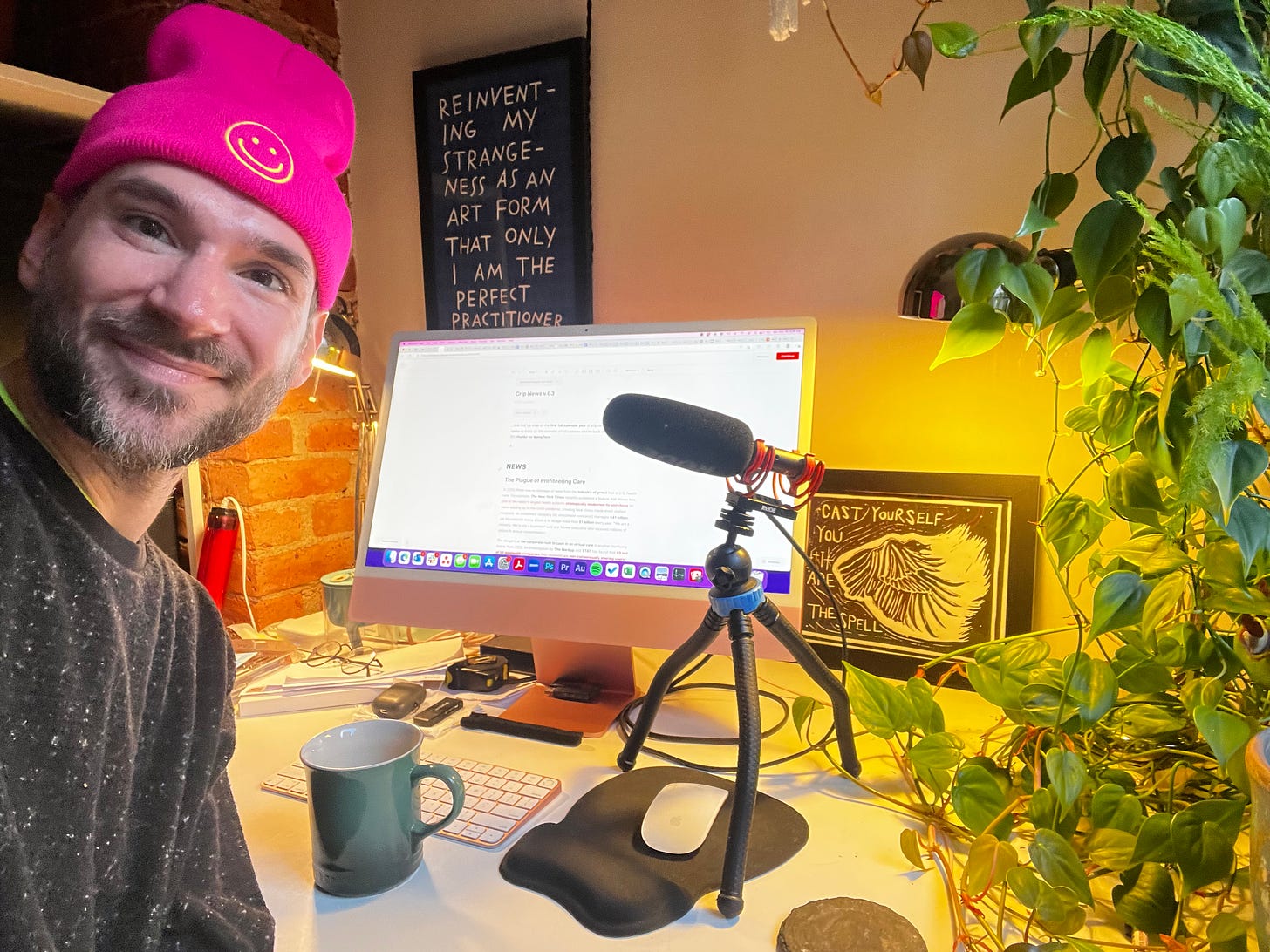 i’m a scruffy white person wearing a bright pink beanie with a neon green smiley face on my forehead. i’m at my desk with an imac and microphone, a heart-shaped philodendron plant sprawling out next to me, a mug for tea, and a few artworks the help enchant the newsroom.