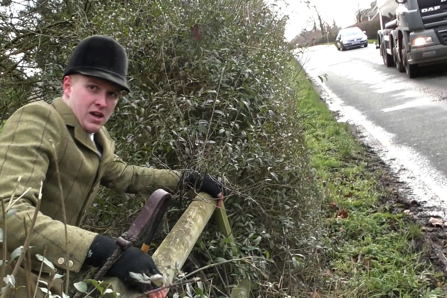 Man in hunting gear stares at the camera as he's about to climb out of undergrowth, over a wooden fence and onto a road with a lorry and car on it.