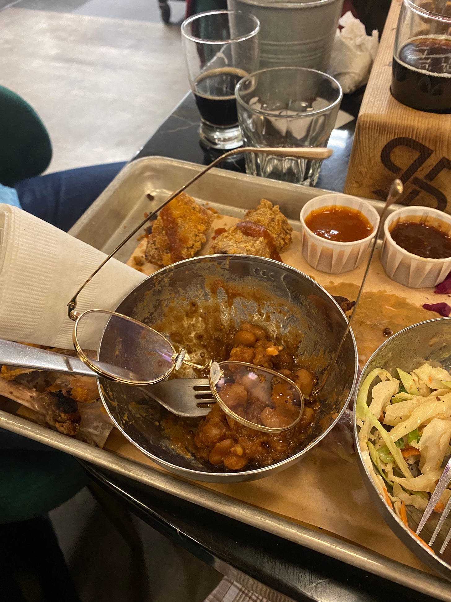 A metal plate of barbecue, partially eaten, with sides in metal bowls and sauces in paper cups. A pair of gold-rimmed glasses have fallen into a bowl of beans. The arms of the glasses stick out and one lens is resting on top of the fork.