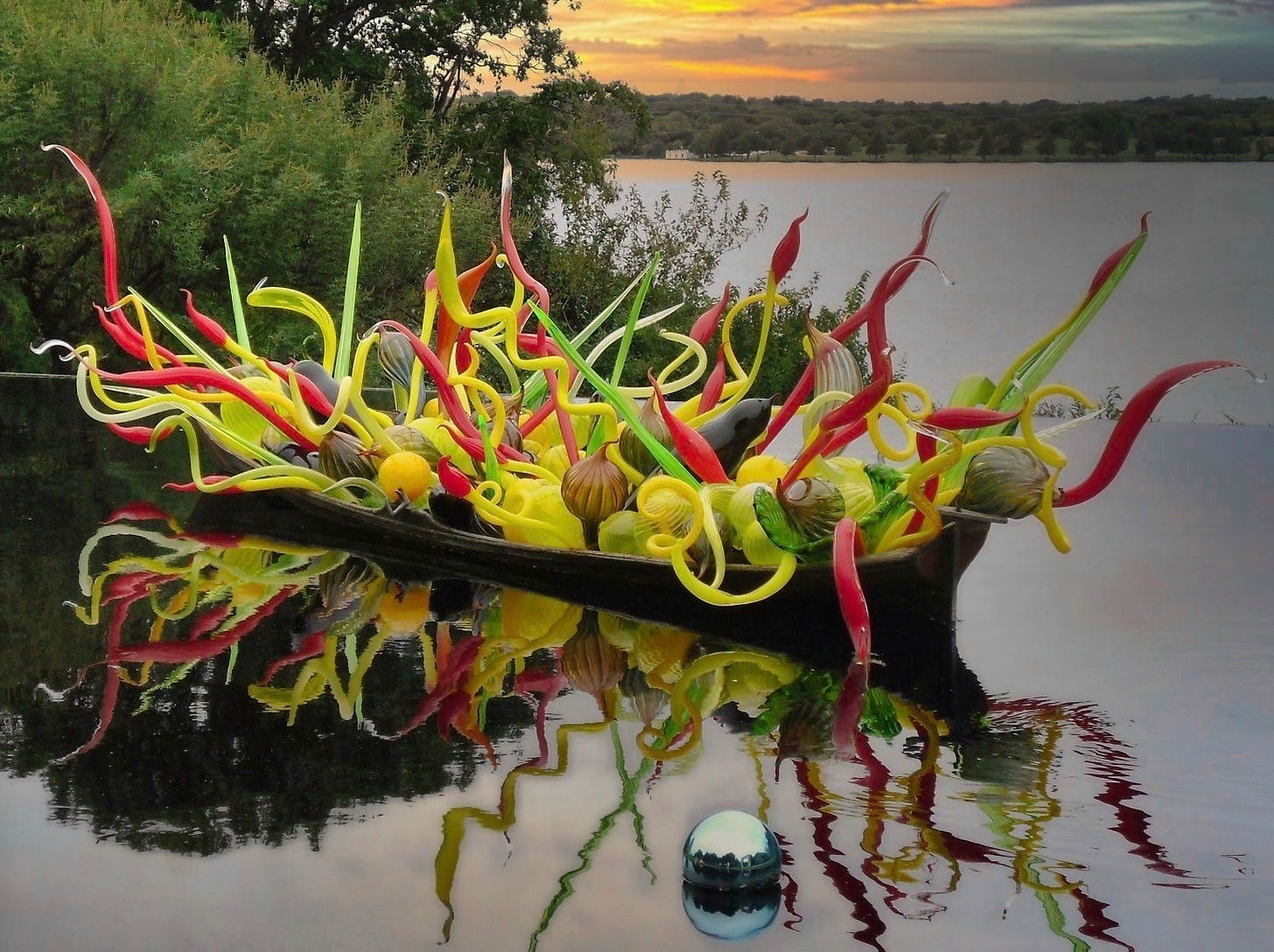A Don Chihuly sculpture of blown glass in orange, yellow, red, lime green in various shaes of globes and long speindles hanging from a flat bottom boat on a lake with the reflection in the foreground and the setting sun behind clouds in the background